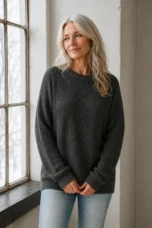 Annabelle charcoal grey waffle-knit sweater worn by a woman with long silver hair and hands loosely clasped beside a window.