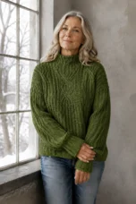 June olive-green cable-knit sweater on the main product model, gently holding her opposite wrist beside a winter window