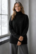 Kathleen black textured turtleneck sweater, model by a window with long wavy hair and hands resting gently across her wrist.