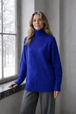 Kathleen dark blue textured turtleneck sweater, model with long wavy hair standing at an angle with one hand on a window ledge.