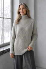 Kathleen grey textured turtleneck sweater, model with long wavy hair looking toward a window and lightly holding the hem.