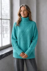 Kathleen light teal textured turtleneck sweater, model with long wavy hair looking toward a window with hands loosely clasped.