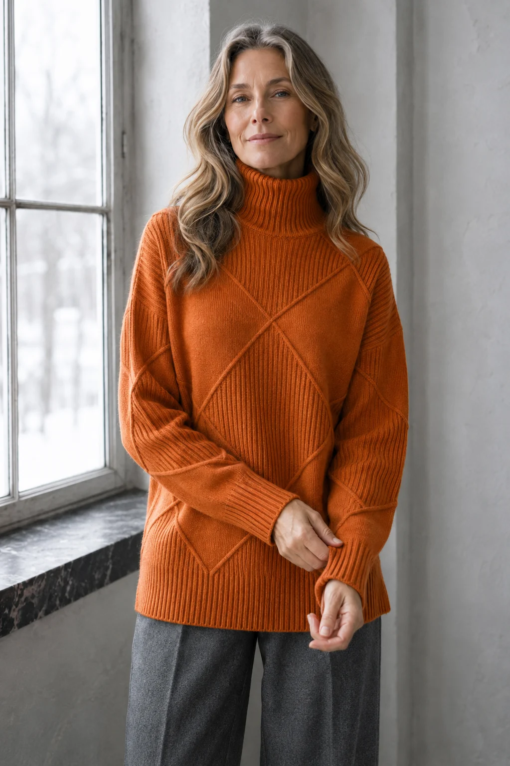Kathleen orange textured turtleneck sweater, model with long wavy hair gently adjusting a cuff by a window.