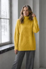 Kathleen yellow textured turtleneck sweater, model with long wavy hair gently touching a strand beside a window.