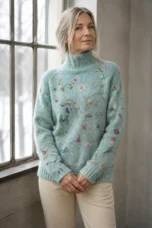 Marcia ice-blue floral turtleneck sweater, worn by a silver-blonde woman with hands loosely clasped beside a window.